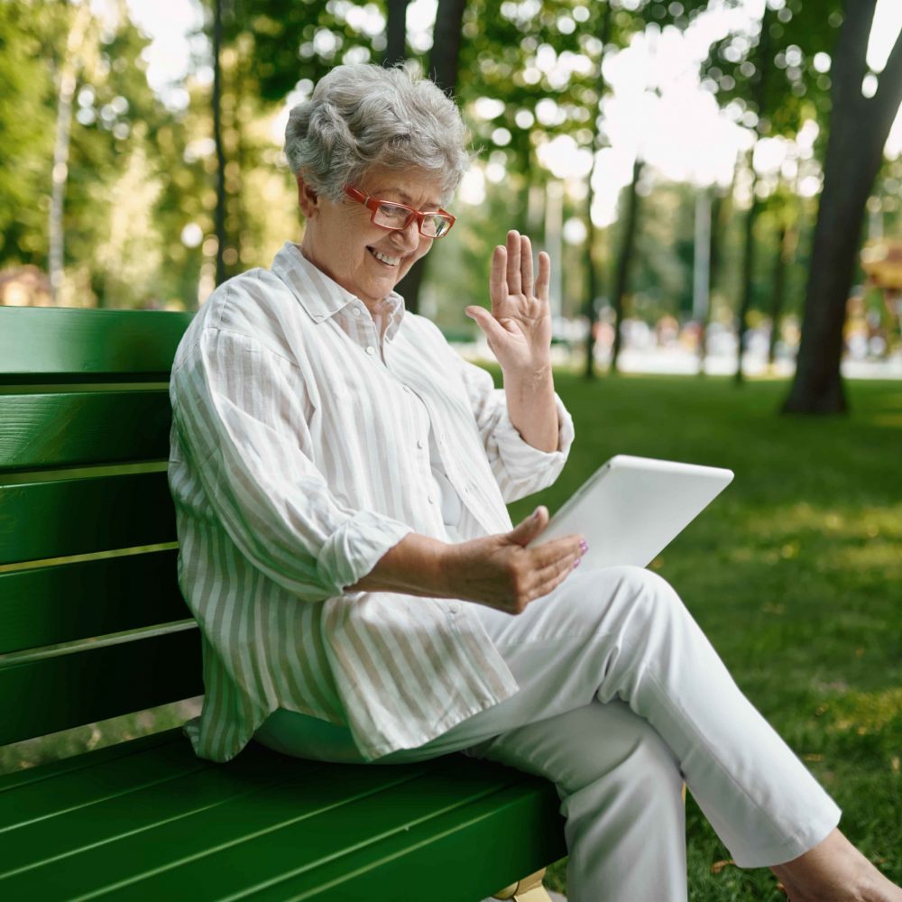 An elderly woman in glasses using laptop on the bench in summer park. Aged people lifestyle. Pretty grandmother having fun outdoors, old female person on nature