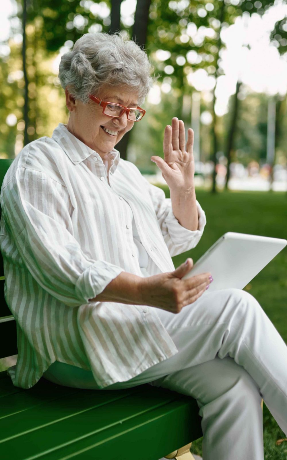 An elderly woman in glasses using laptop on the bench in summer park. Aged people lifestyle. Pretty grandmother having fun outdoors, old female person on nature